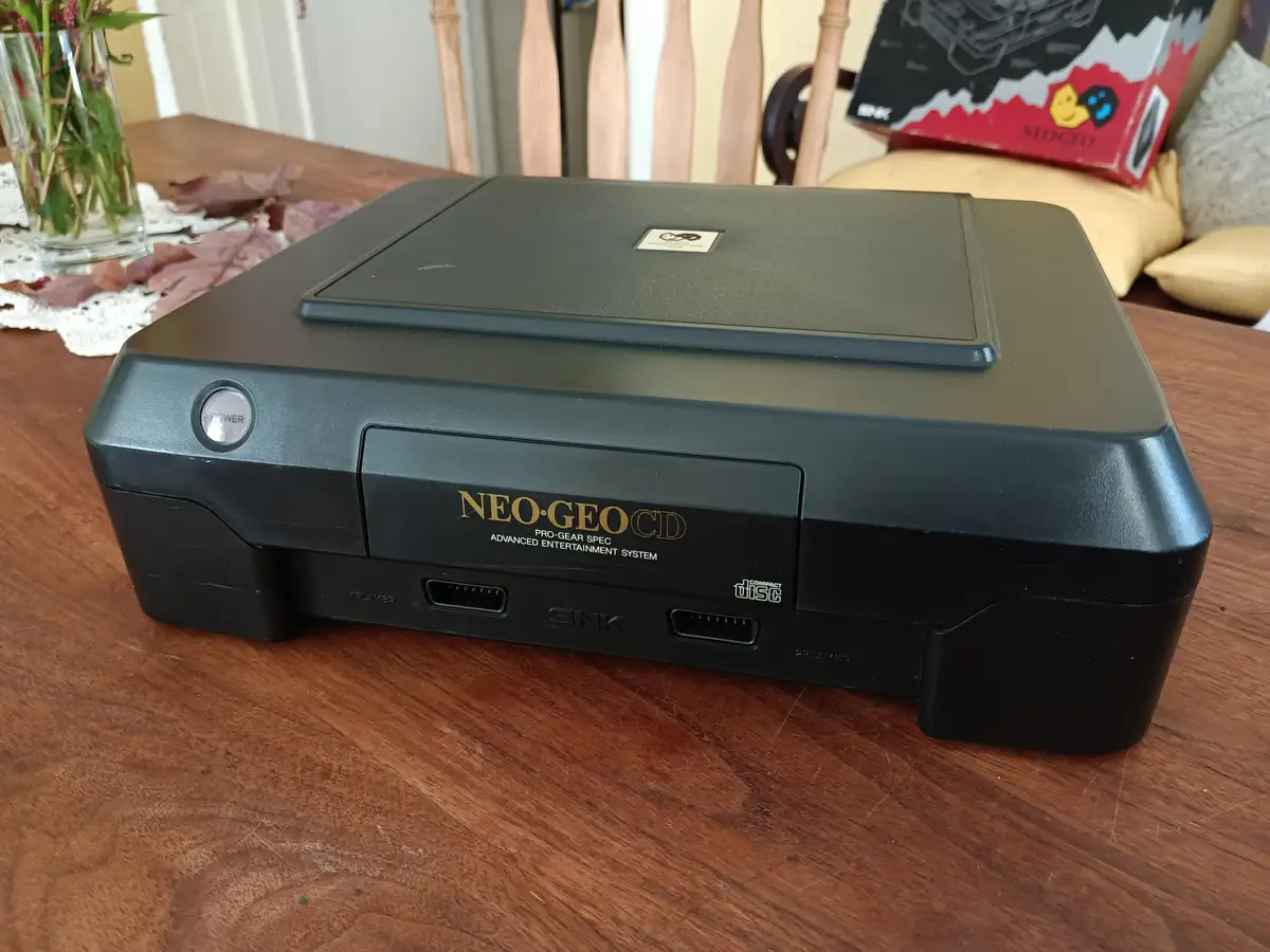 A black SNK Neo-Geo CD console sits on a wooden table, with its front panel showing the 'NEO-GEO CD' logo, 'PRO-GEAR SPEC'…