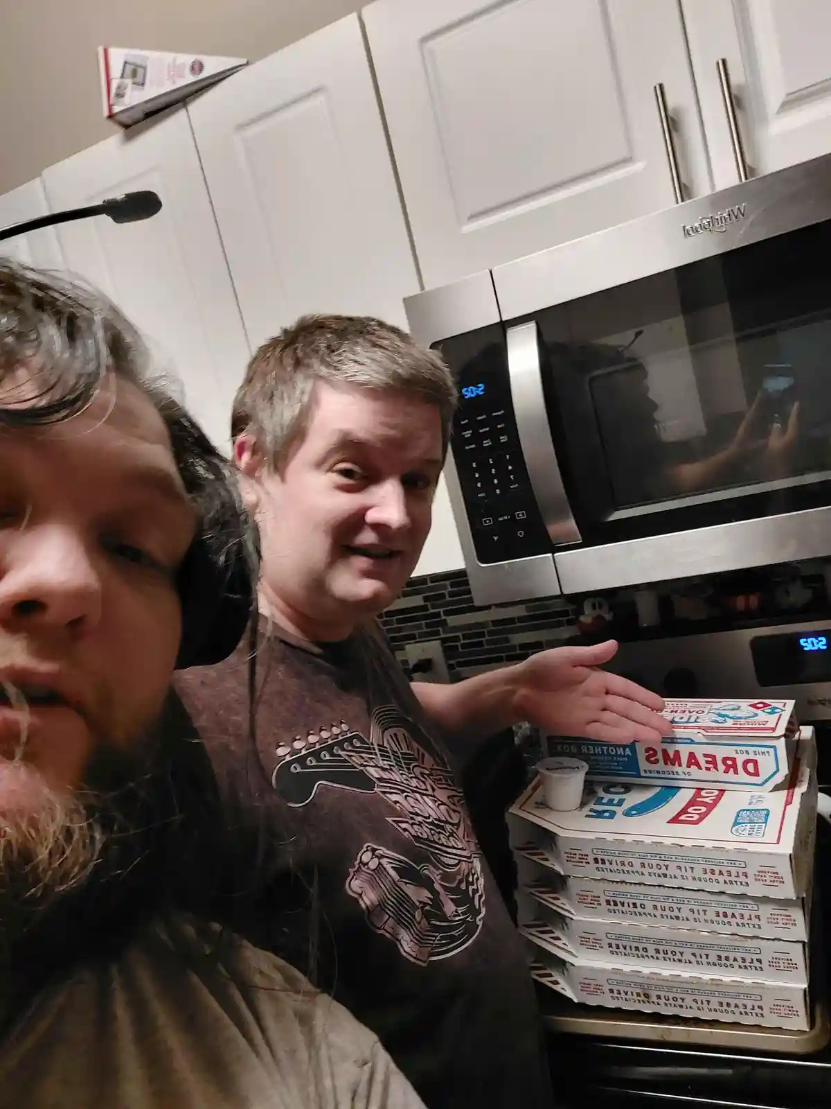 Two men are in a kitchen, one taking a selfie while the other gestures toward a tall stack of pizza boxes. The top box is…