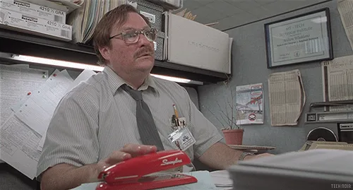 A man in a gray shirt and tie sits at a cluttered office desk, holding a red stapler. He looks upward with a skeptical…