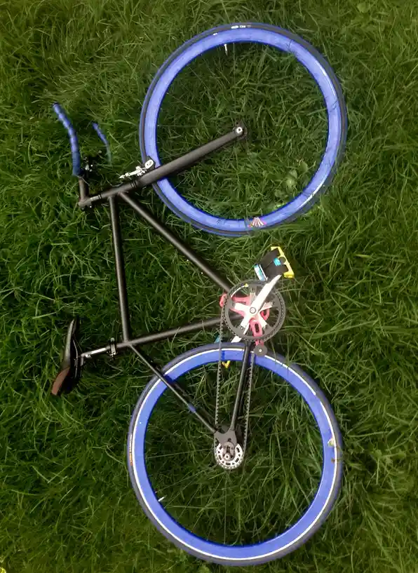 An overhead shot of a black fixed-gear bicycle with bright blue wheels lying on green grass. The bike's crankset is replaced…