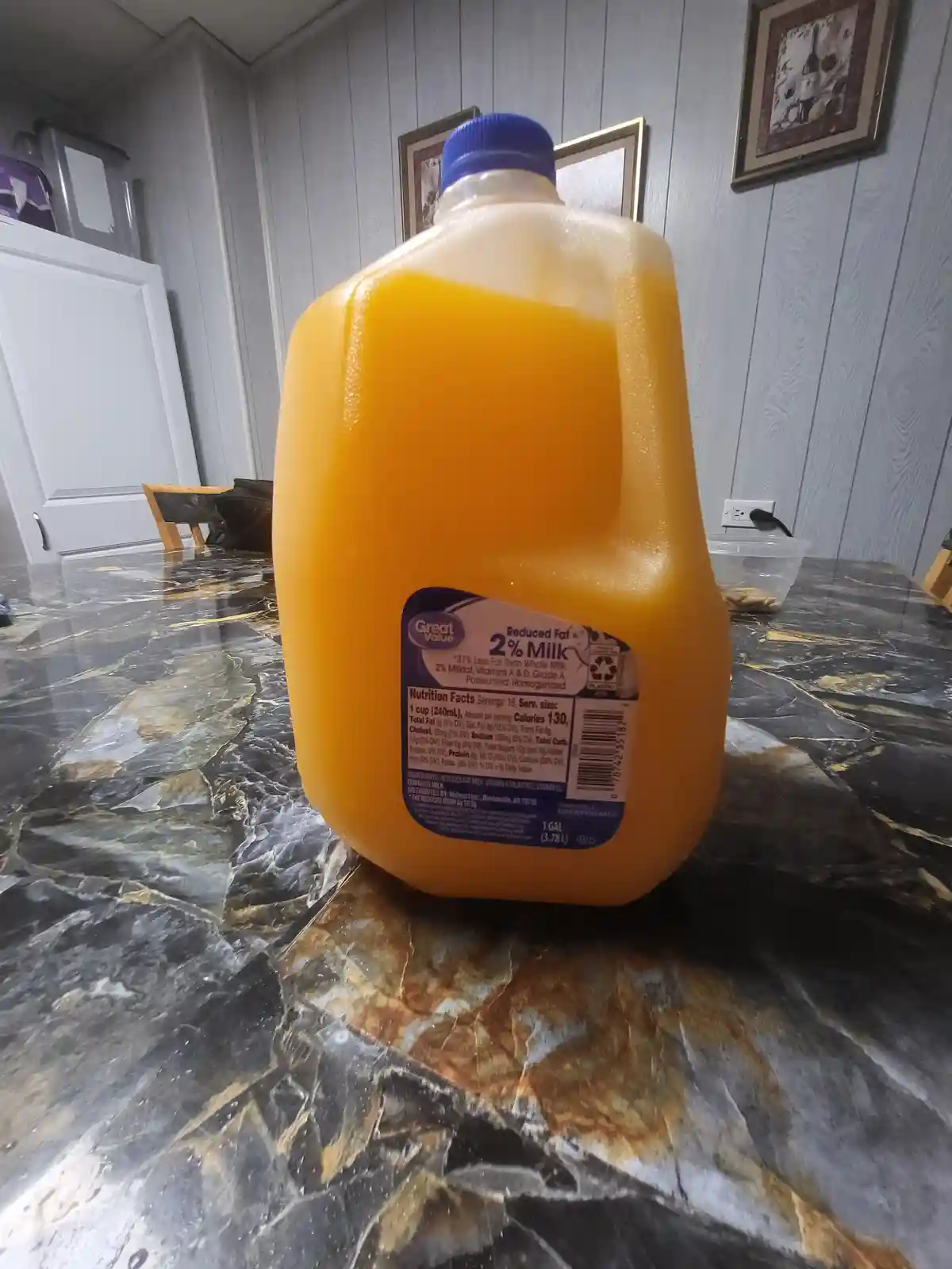 A large, one-gallon plastic jug of Great Value brand 2% reduced fat milk sits on a dark, marbled kitchen countertop. The…