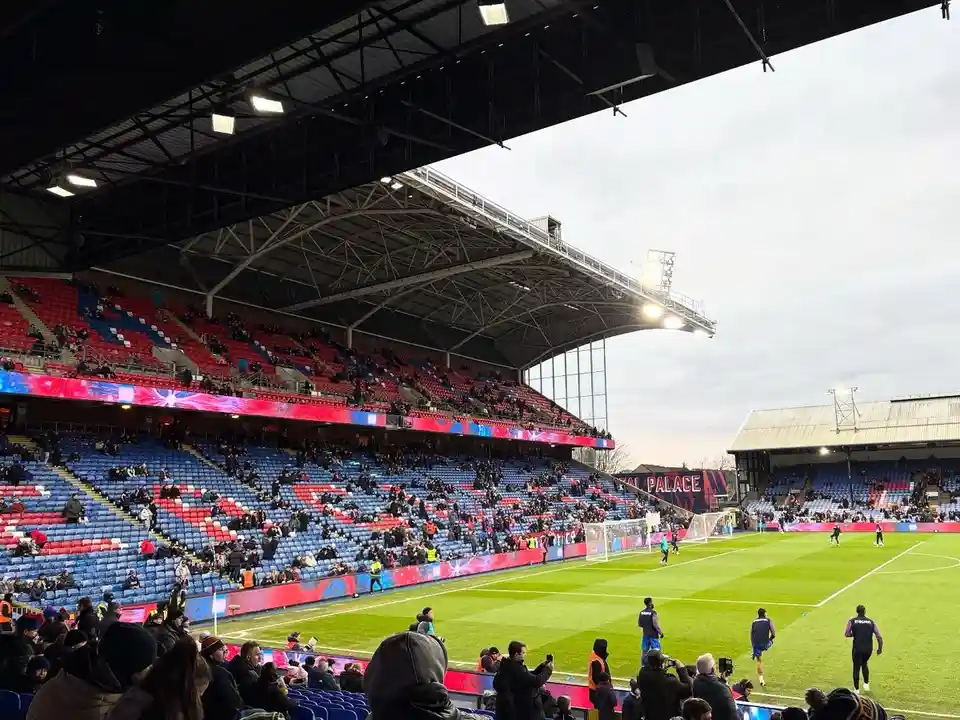 A wide shot of a football stadium with partially filled red and blue seats under an overcast sky. Players are on the green…
