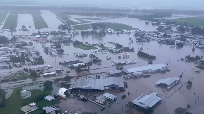 an aerial view of a flooded rural area with houses, barns, and roads submerged in murky water under an overcast sky…