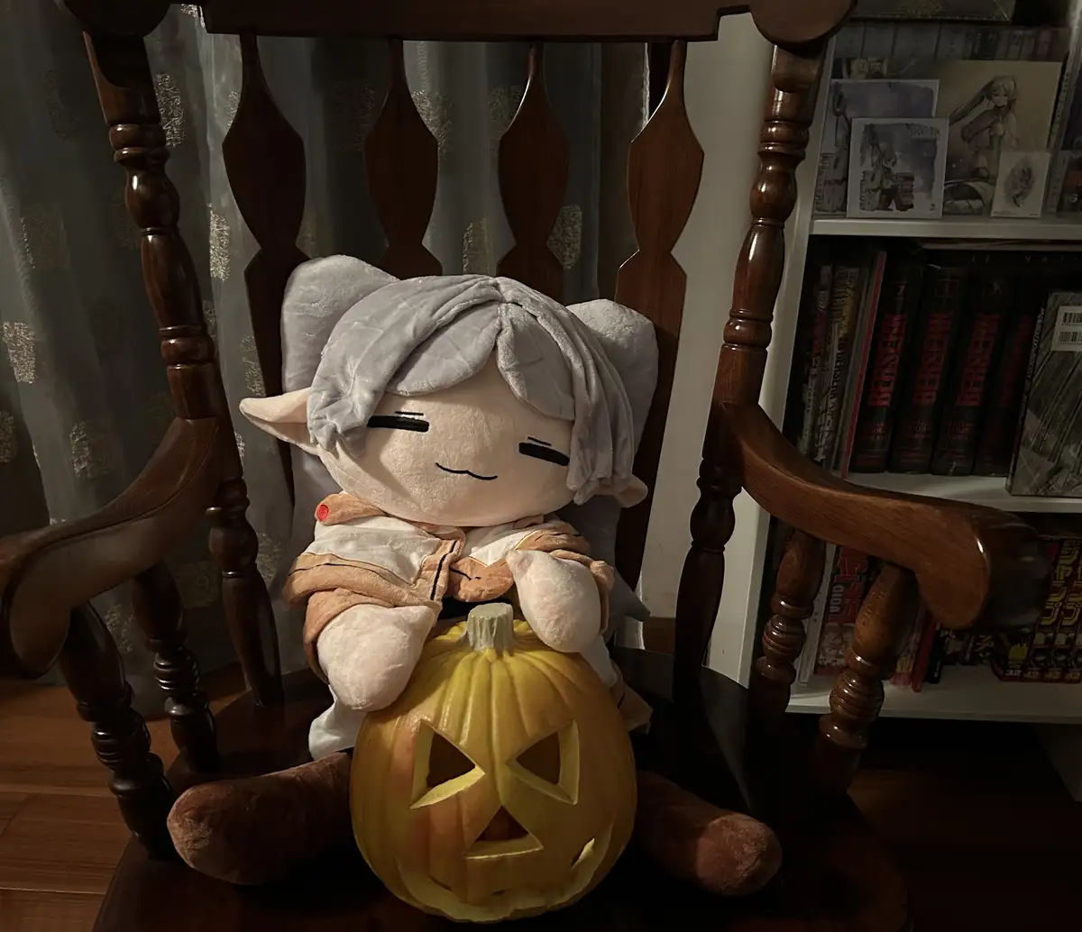 A plushie of a character with grey hair and pointed ears sits in a wooden rocking chair, holding a carved jack-o'-lantern…