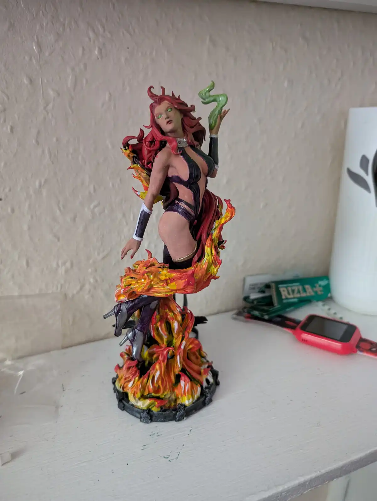A painted figurine of a female comic book character with red hair, green eyes, and fiery orange-yellow flames around her…