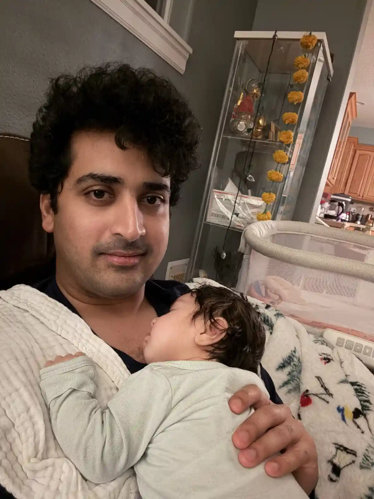 A man with dark curly hair holds a sleeping baby wrapped in a white blanket, looking directly at the camera with a serious…