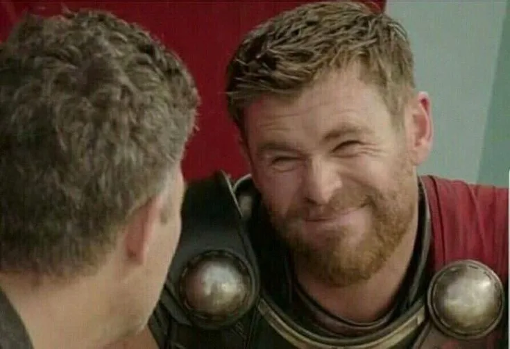 a close-up of actor Chris Hemsworth as Thor, smiling smugly while looking at another person whose back is to the camera…