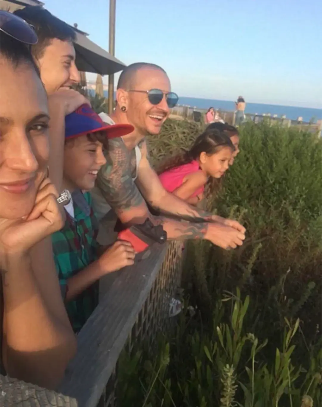 A group of people, including a man with tattoos and sunglasses, a woman, and two children, are leaning over a wooden railing…