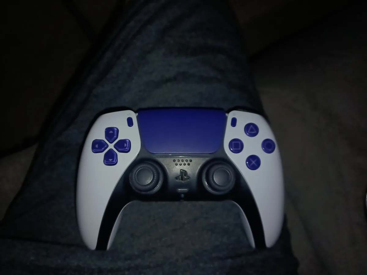 A white and blue PlayStation 5 DualSense controller rests on a person's dark gray pants in a dimly lit room. The controller…