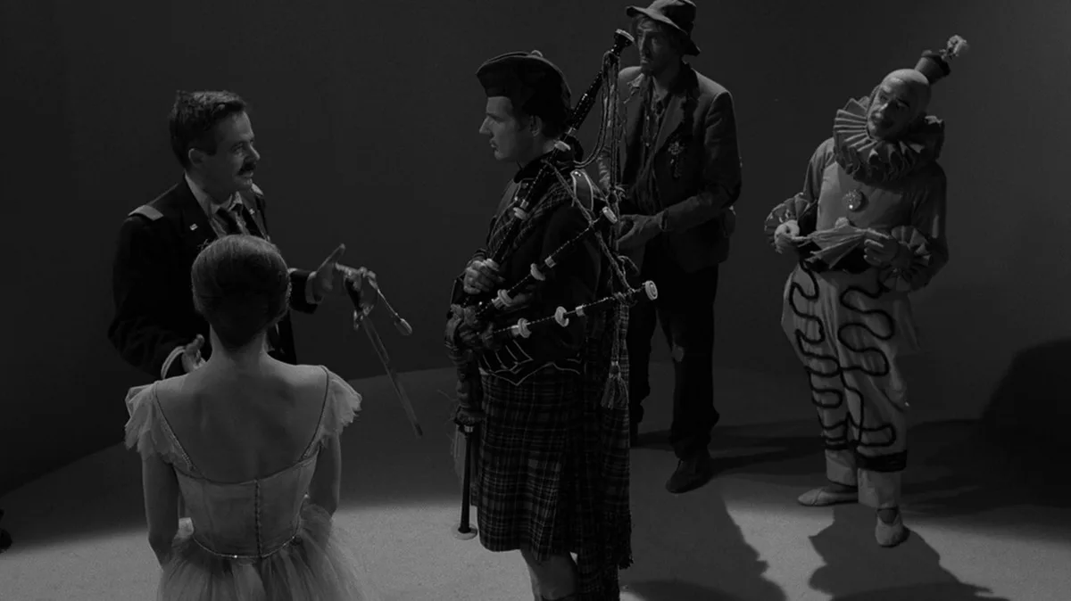 A black-and-white scene from a film shows a man in a military-style uniform gesturing toward a ballerina in a tutu, while…