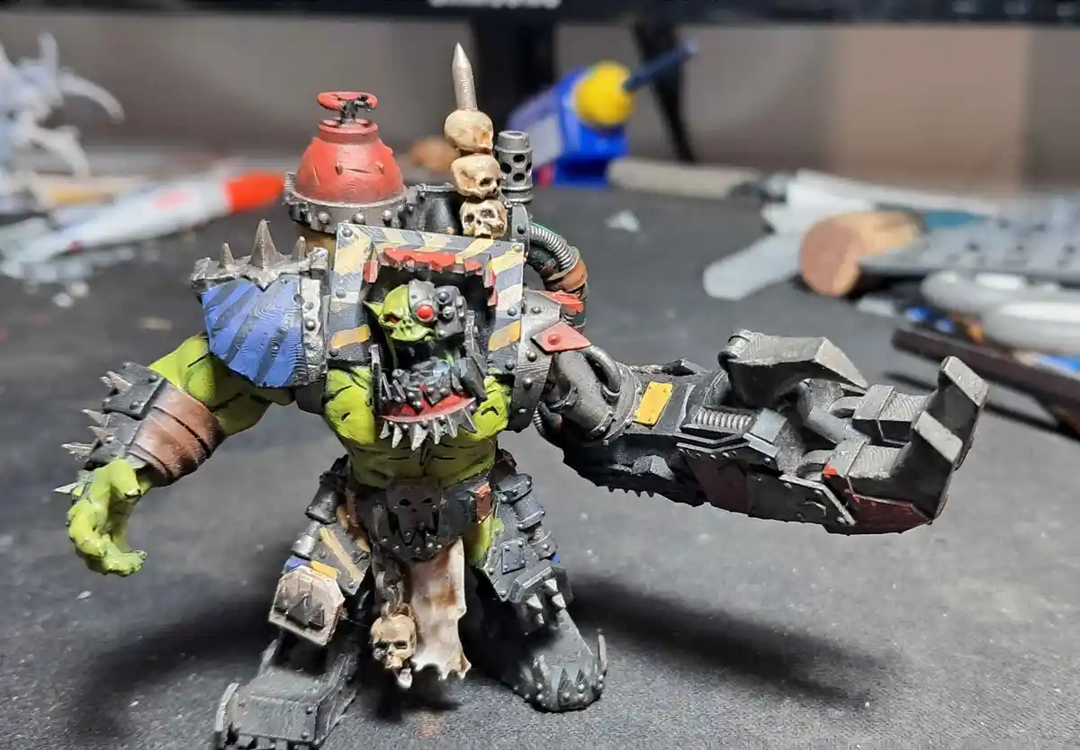 A heavily armored, green-skinned fantasy miniature with a spiked helmet, skulls, and a massive mechanical arm, standing on a…