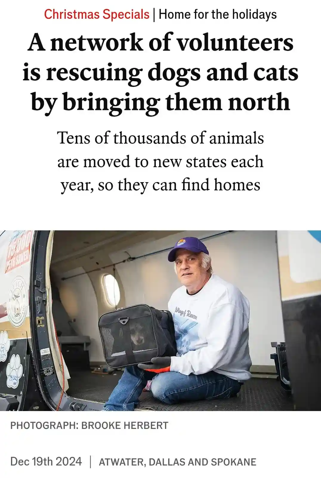 A man in a white sweatshirt and blue cap sits inside a converted airplane, holding a pet carrier with a dog inside. The…