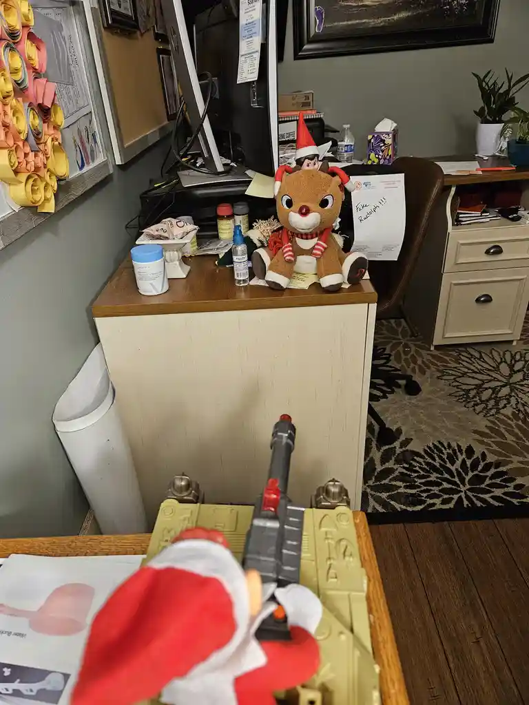 A first-person view from a toy tank with a Santa hat, aiming a toy gun at a plush reindeer toy on a cluttered office desk. A…