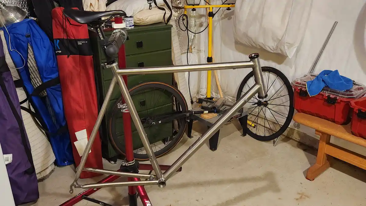 A shiny, custom-made bicycle frame is mounted on a red repair stand in a cluttered garage, surrounded by tools, storage…