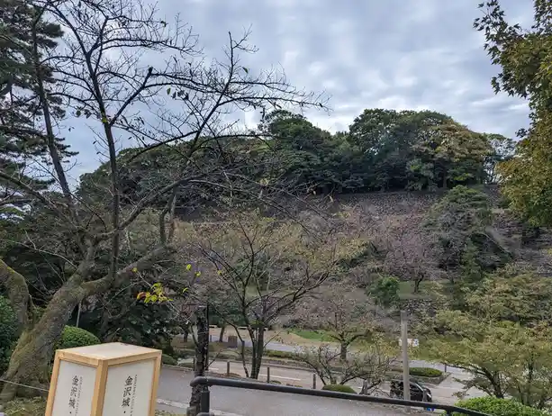 A photo of Kanazawa Castle's stone walls and surrounding trees under an overcast sky, with a wooden sign in the foreground…