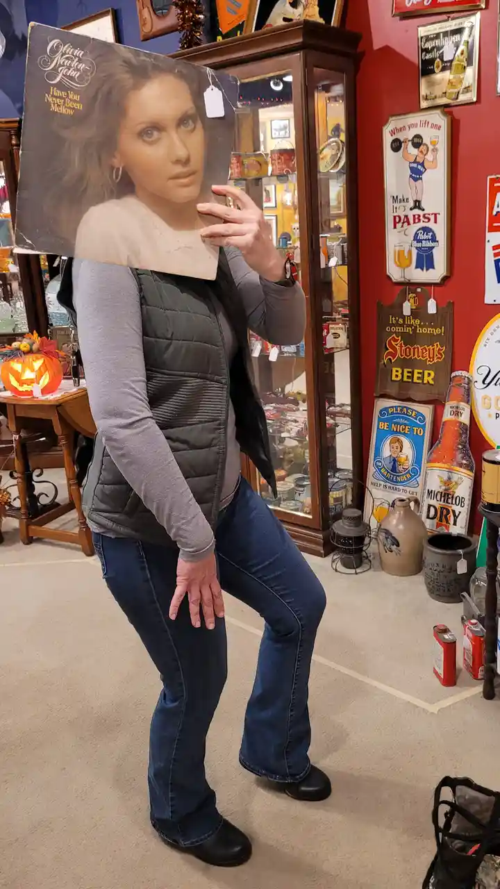 A person in a grey shirt, black vest, and jeans holds up a large Olivia Newton-John album cover titled 'Have You Never Been…