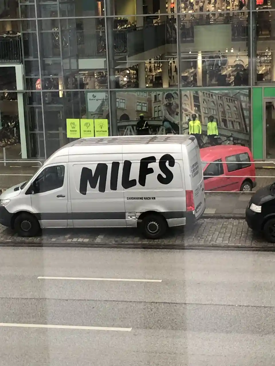 A silver van parked on a city street has the word 'MILFS' spray-painted in large black letters on its side, directly…