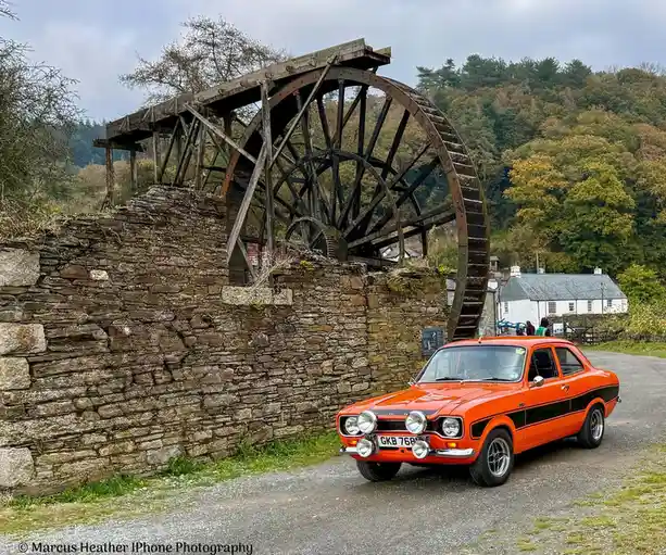 A bright orange vintage Ford Escort with a black stripe is parked on a gravel road in front of a large, weathered wooden…