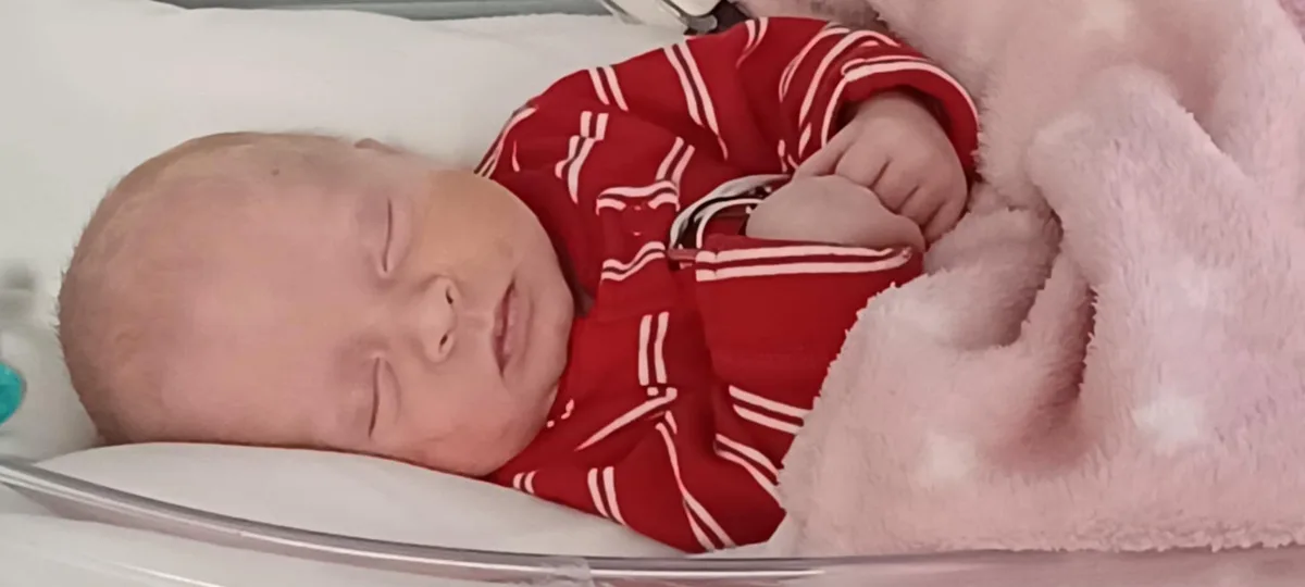 A newborn baby, wearing a red and white striped onesie, sleeps peacefully in a clear hospital bassinet, partially covered by…