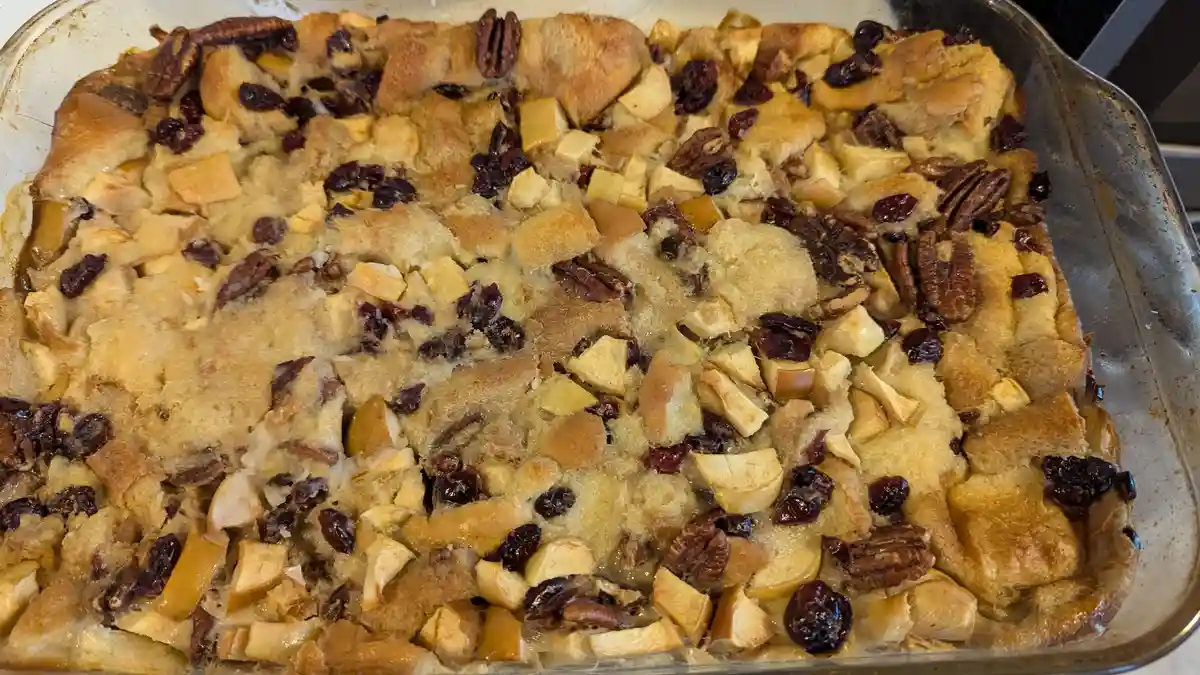 A close-up photo shows a baked dish in a glass baking pan, featuring golden-brown bread cubes mixed with chunks of apple…