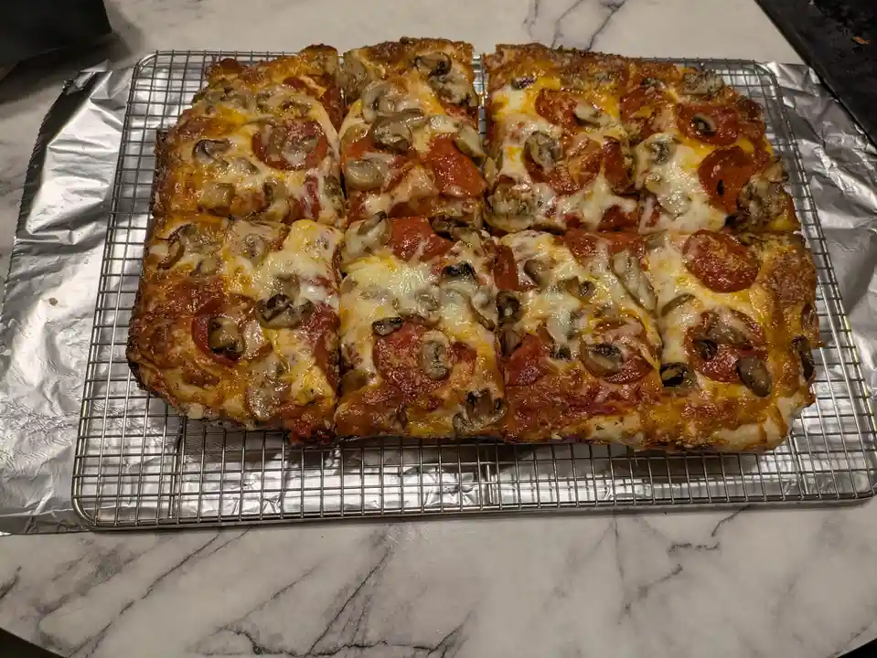 A rectangular pepperoni and mushroom pizza, cut into 8 square slices, rests on a metal cooling rack atop aluminum foil on a…