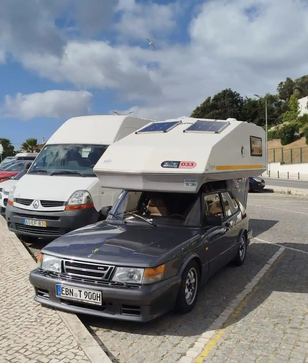 A dark gray Saab 900 sedan parked in a cobblestone lot with a large white camper shell mounted on its roof, featuring solar…