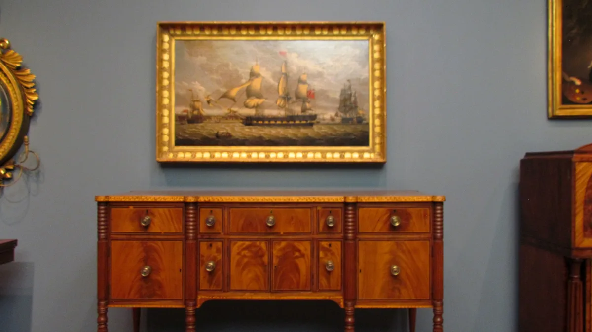 A wooden sideboard with ornate drawers and brass knobs sits beneath a large, gold-framed painting of sailing ships at sea…