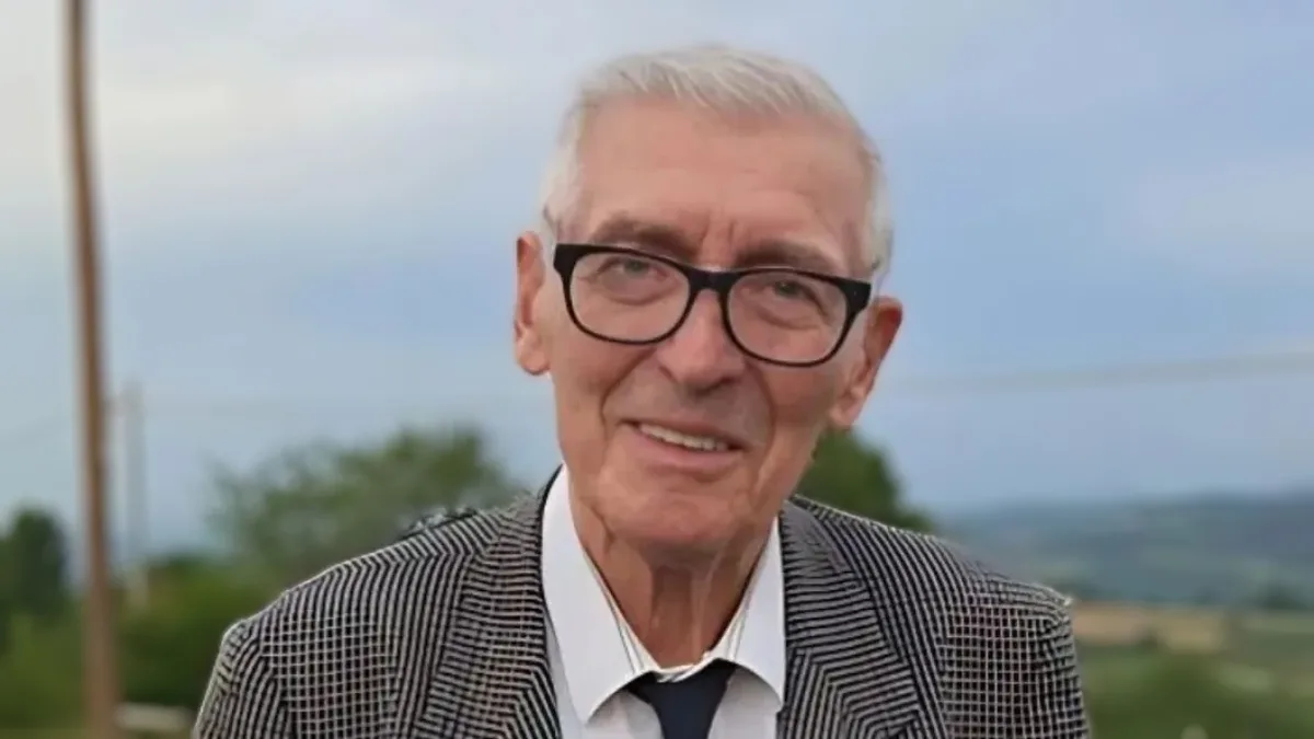 an older man with white hair and black-rimmed glasses smiles at the camera while wearing a checkered blazer, white shirt…