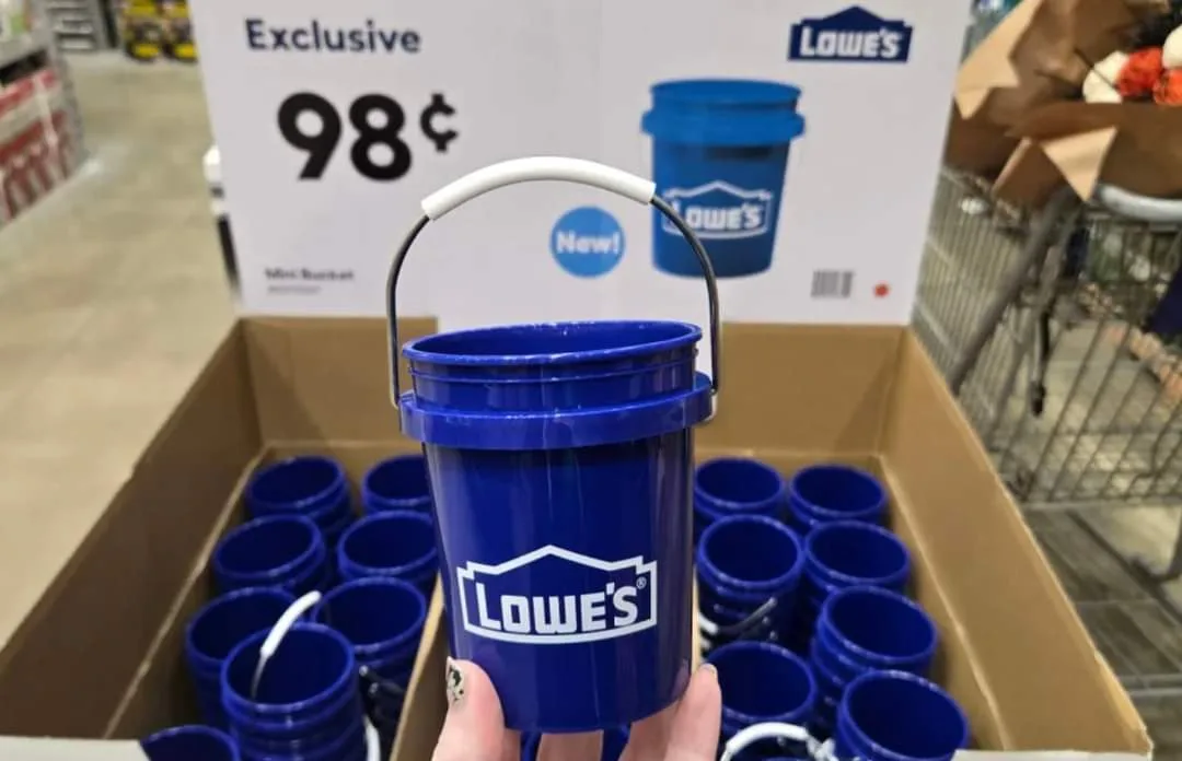 A hand holds a small blue plastic bucket with the Lowe's logo, in front of a display box filled with identical buckets. A…