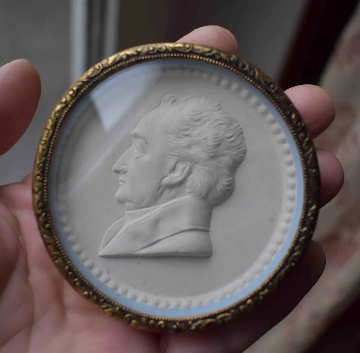 A hand holds a circular, ornate gold-framed medallion containing a white relief sculpture of a man's profile. The text…