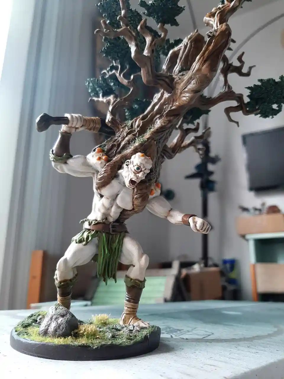 A painted miniature figurine of a muscular humanoid creature with a large, gnarled tree growing out of its back, wielding a…