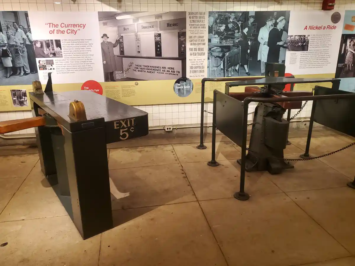 An old-fashioned subway turnstile with 'EXIT 5¢' written on its side is displayed in a museum exhibit, surrounded by…
