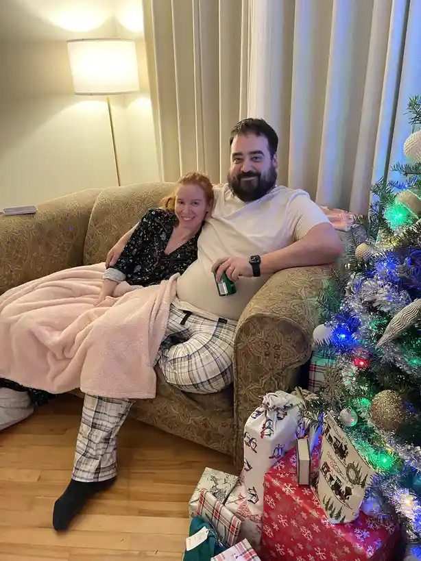 A man and woman in pajamas sit cozily on a couch next to a lit Christmas tree with wrapped gifts, the man holding a can and…