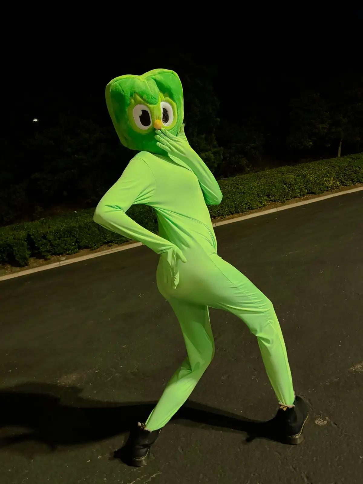 A person in a full-body, bright green costume resembling the character Jujubee from 'The Owl House' stands on a dark asphalt…