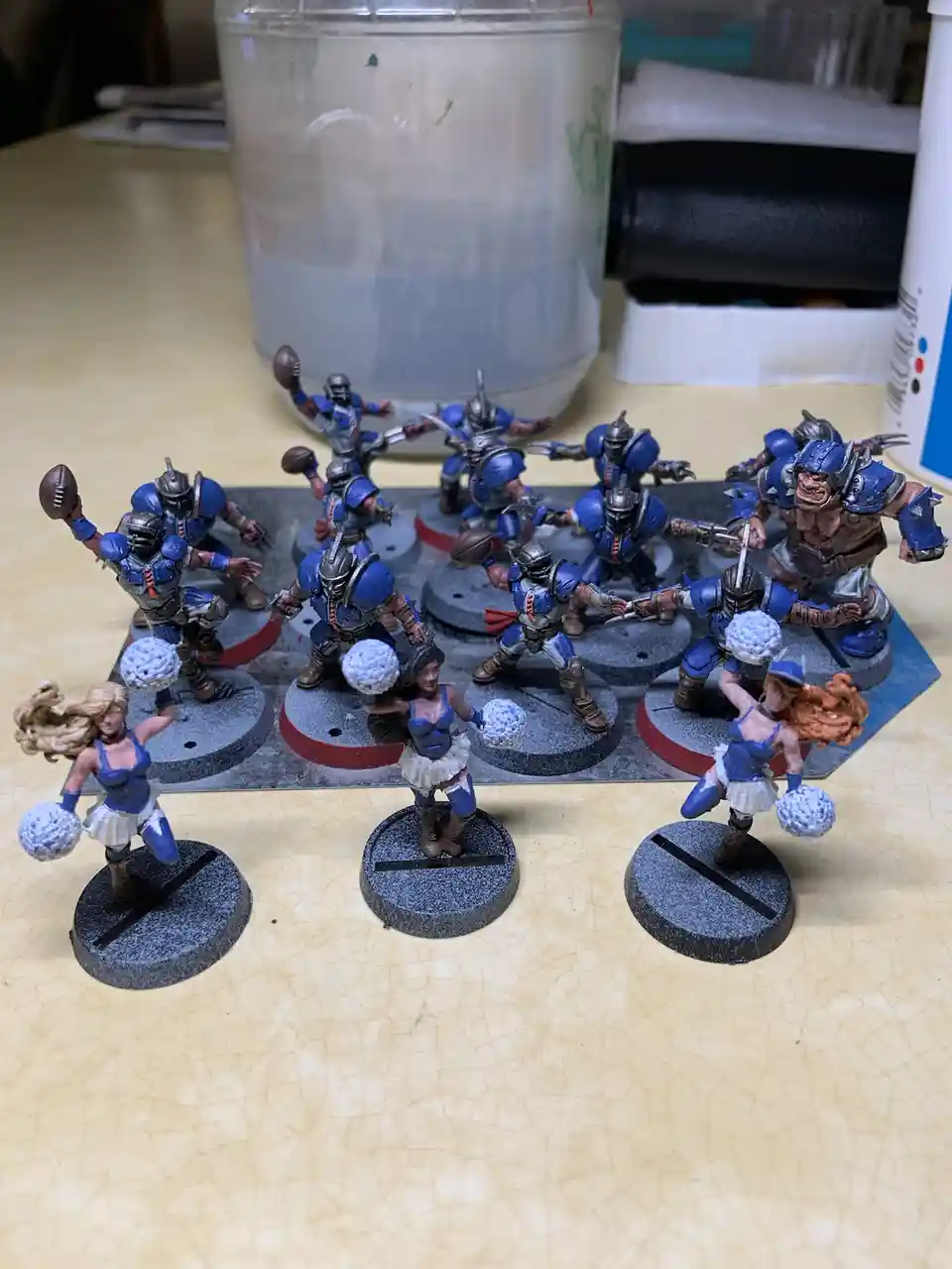 A collection of painted miniature figurines, mostly armored warriors in blue, are arranged on a tabletop. Three cheerleader…