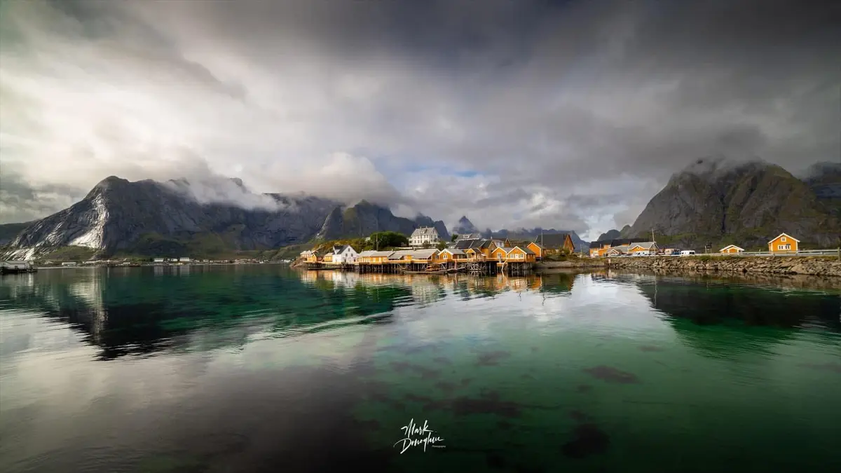 A scenic photograph of a coastal village with colorful wooden houses on stilts over calm, reflective water, surrounded by…