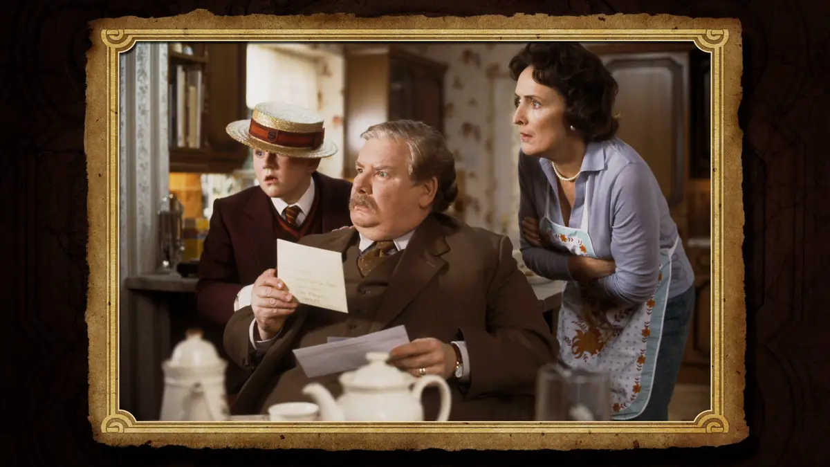 A still from a period drama shows a man in a brown suit holding a letter, looking shocked, with a young boy in a straw hat…