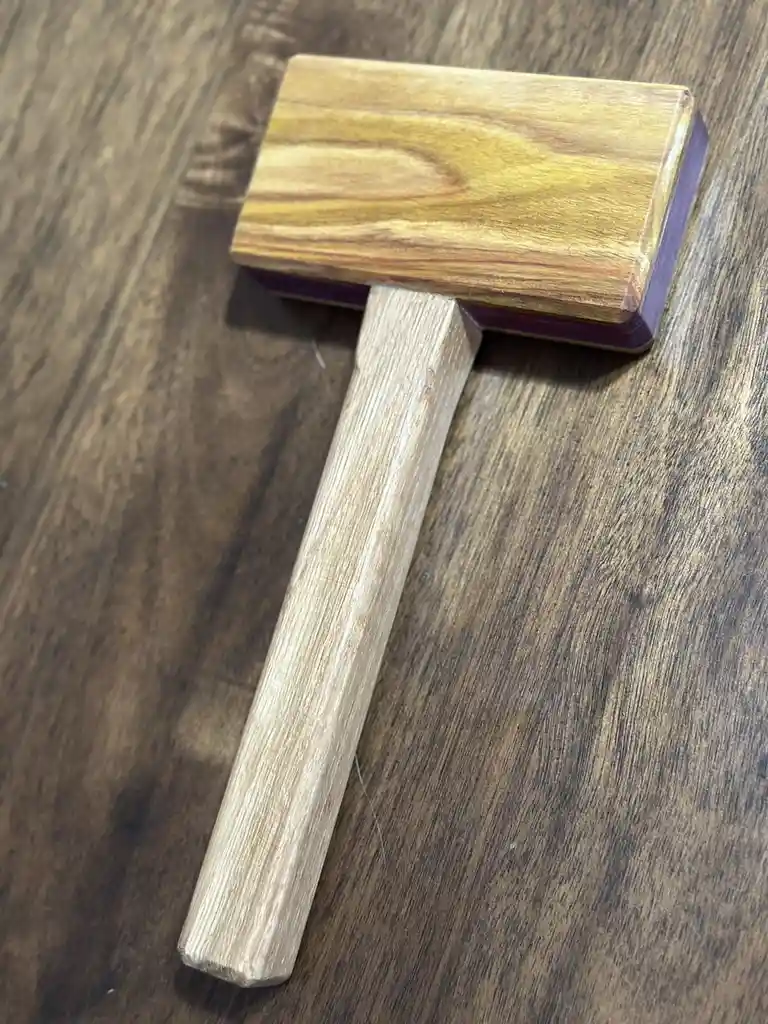 A wooden mallet with a light-colored handle and a rectangular head made of layered wood, one layer showing a purple hue…