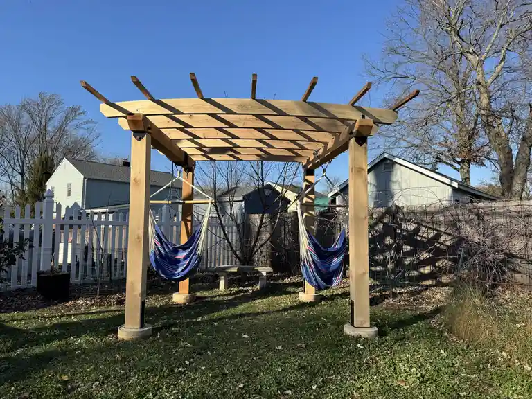 A wooden pergola stands in a backyard with two blue striped hammocks hanging from its beams, set against a backdrop of a…