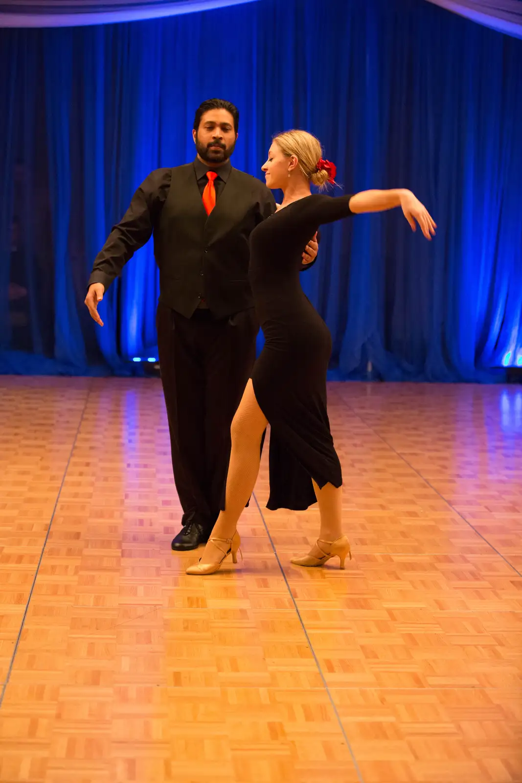 A man and woman in formal dance attire are mid-dance pose on a wooden ballroom floor with blue curtains in the background…