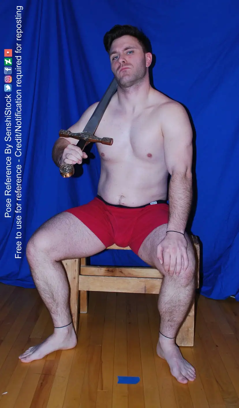 A shirtless man with dark hair and a beard sits on a wooden chair against a blue backdrop, holding a large ornate sword…