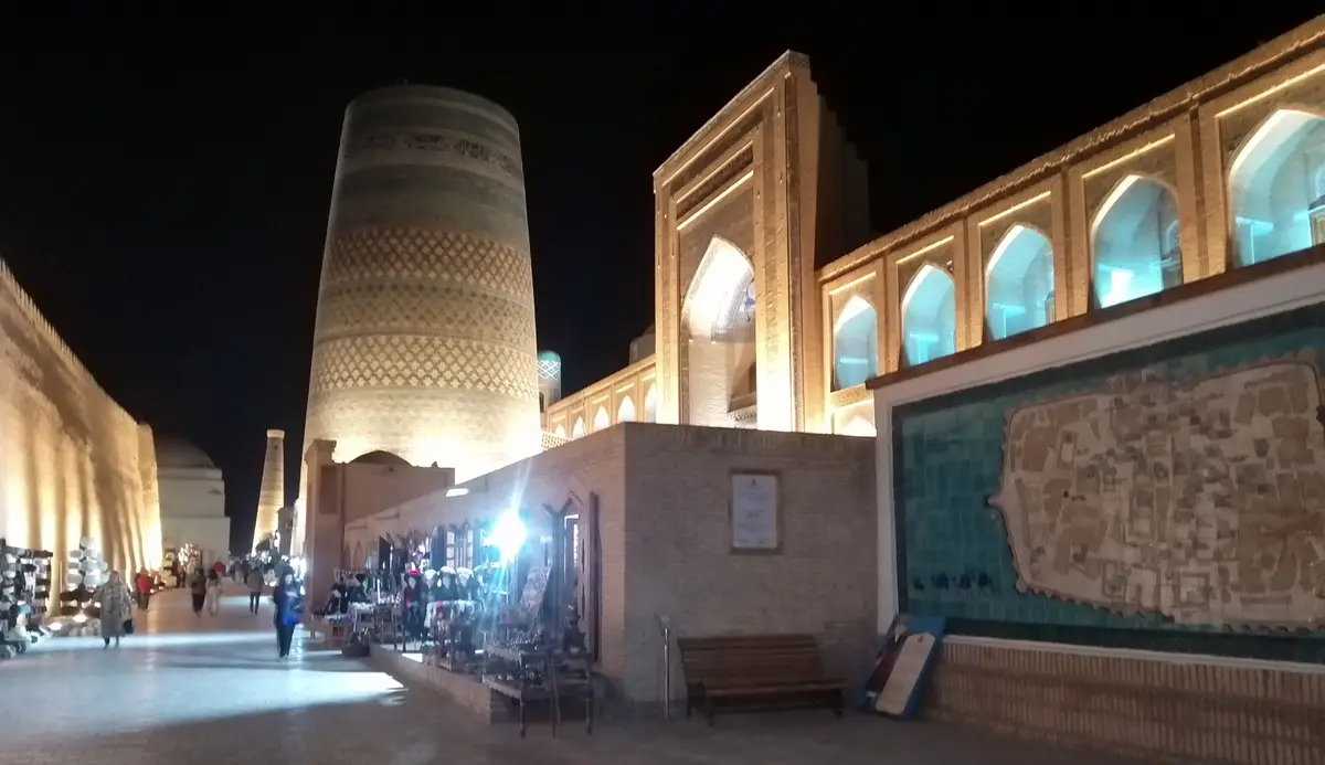 A nighttime photo of a historic, illuminated Islamic architectural complex with a tall cylindrical tower and arched…
