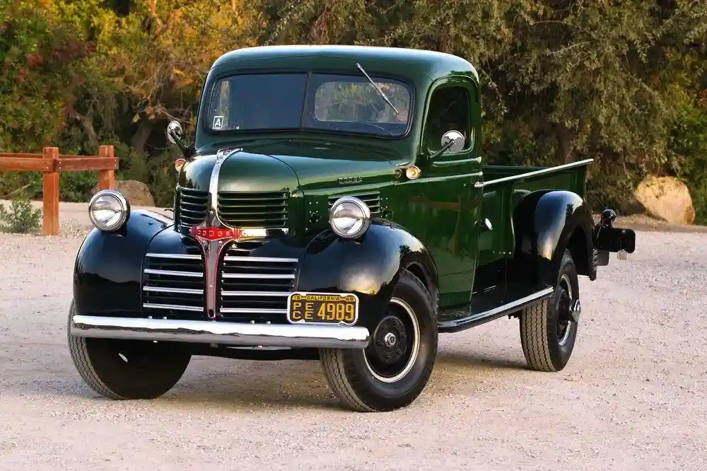 A vintage green 1940s Dodge pickup truck is parked on gravel with trees in the background. The truck has a classic design…