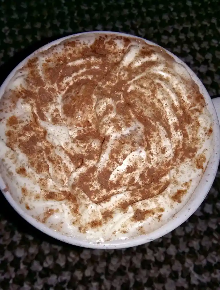 A top-down photo of a white mug filled with a frothy beverage, likely a latte or cappuccino, topped with swirls of white…