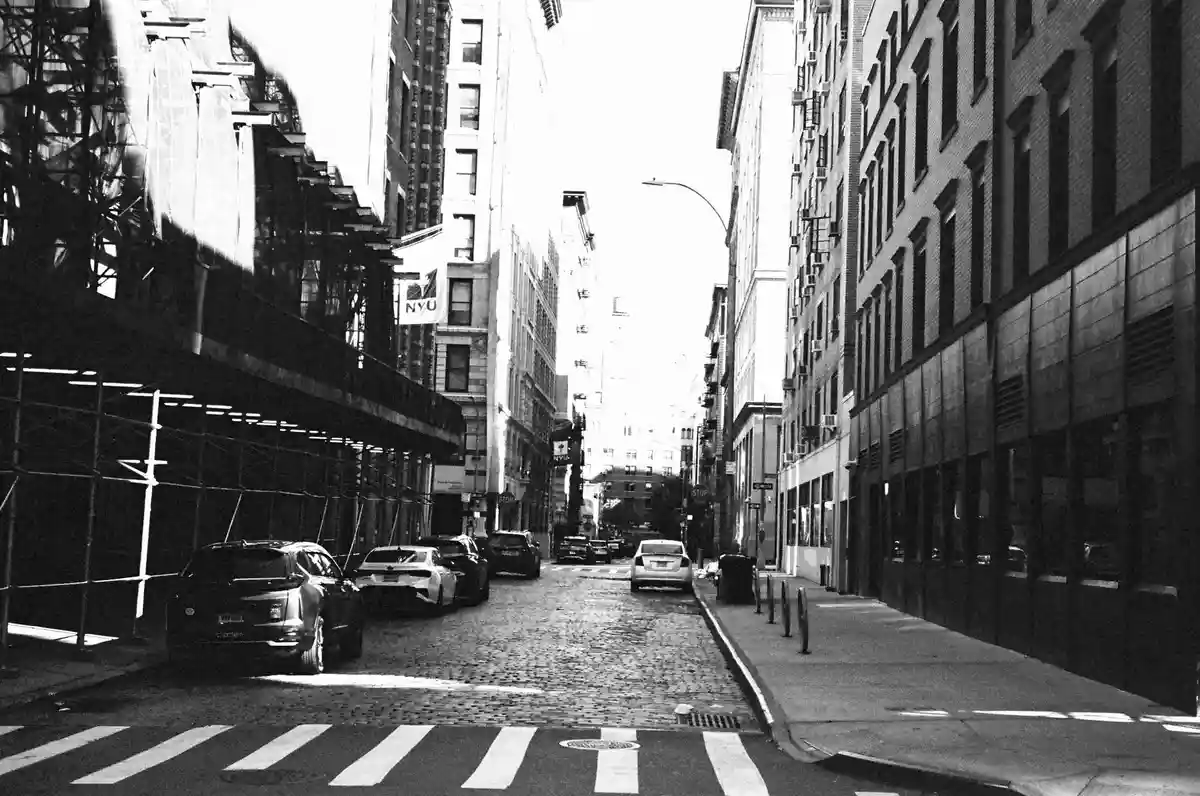 A black-and-white photo of a narrow cobblestone street in a city, flanked by tall buildings with scaffolding on the left and…