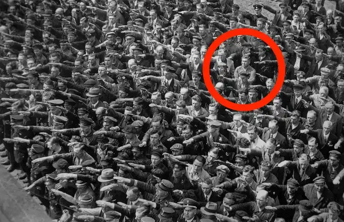 a black and white historical photograph shows a large crowd of men in uniform performing the Nazi salute. One man in the…