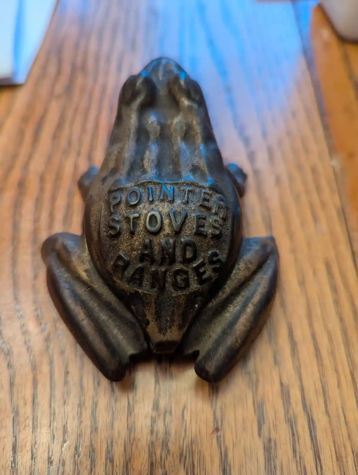 A close-up photo shows a vintage, dark bronze frog-shaped stove knob resting on a wooden surface. The frog's belly is…