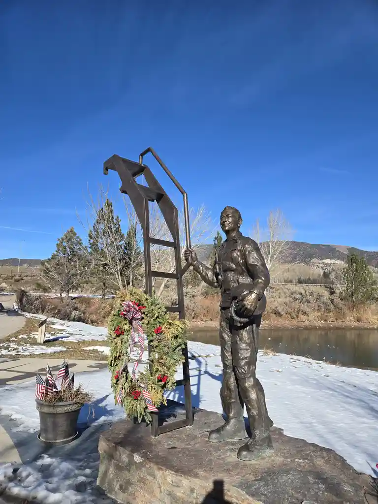 A bronze statue of a smiling firefighter in uniform stands beside a ladder, with a patriotic wreath and American flags…