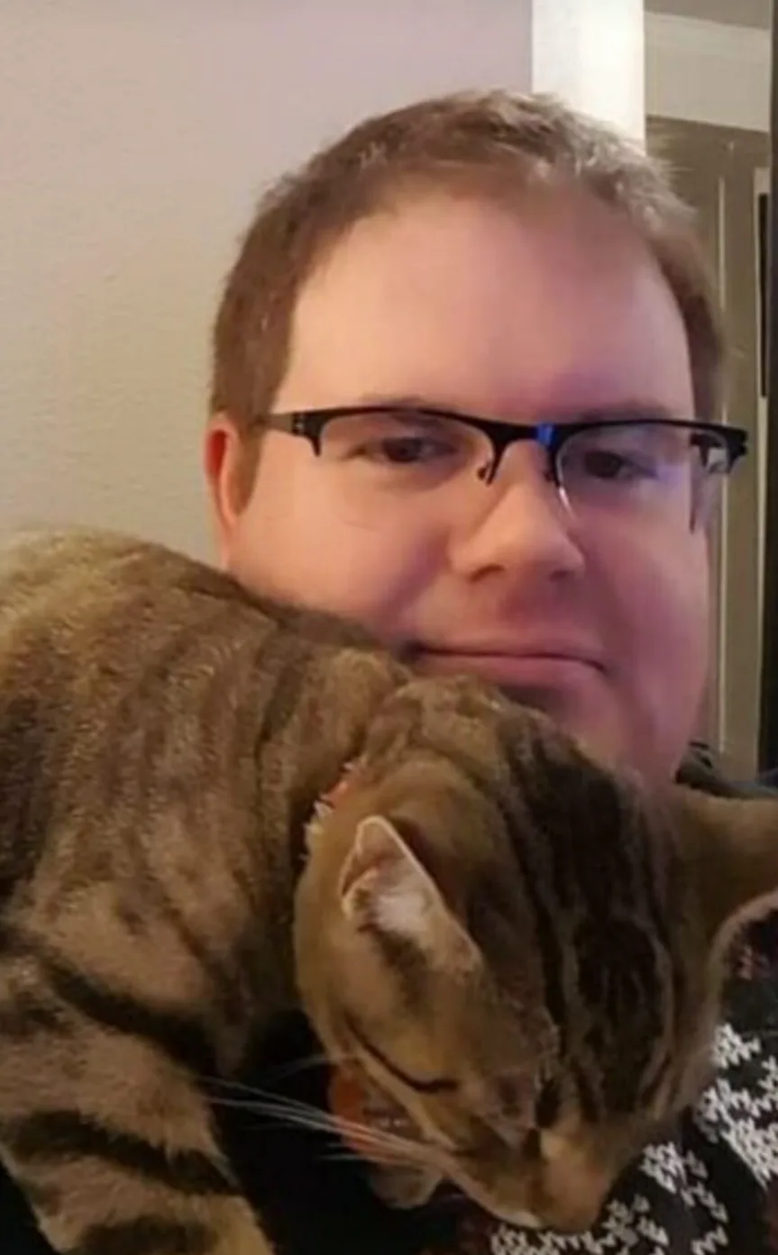 A man with glasses is holding a tabby cat that is resting its head on his shoulder. The cat appears relaxed and content…
