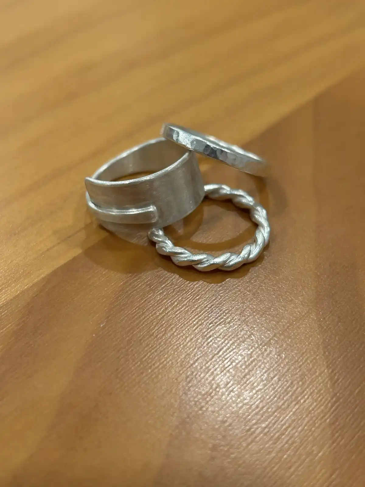 Three silver rings — one thick band, one twisted rope-style, and one flat hammered band — are arranged on a wooden surface…